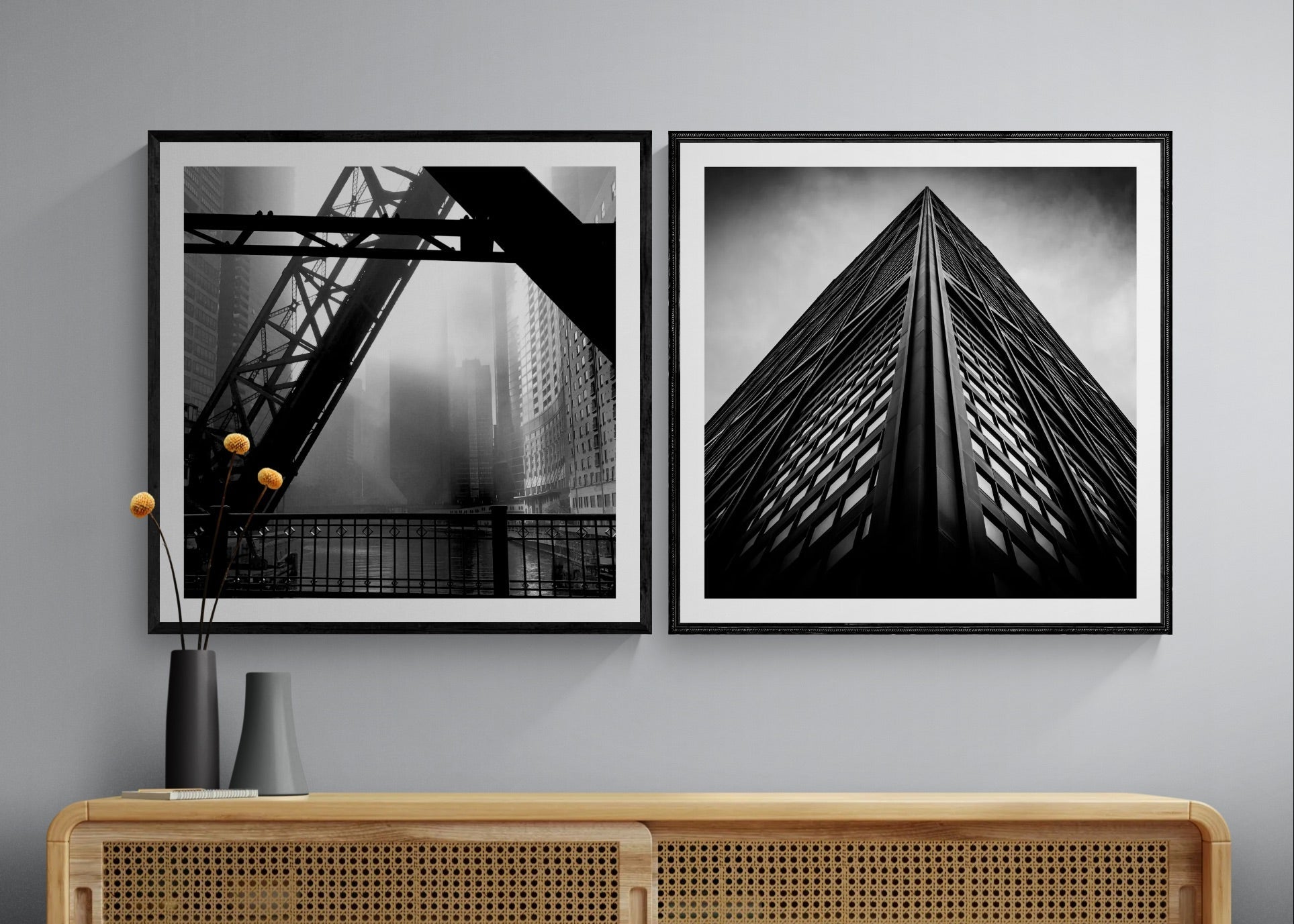 Two framed black and white architectural art prints hang on a light gray wall above a light wood console cabinet with woven front panels. The left print shows a bridge over water, and the right print shows a tall building facade viewed from below.