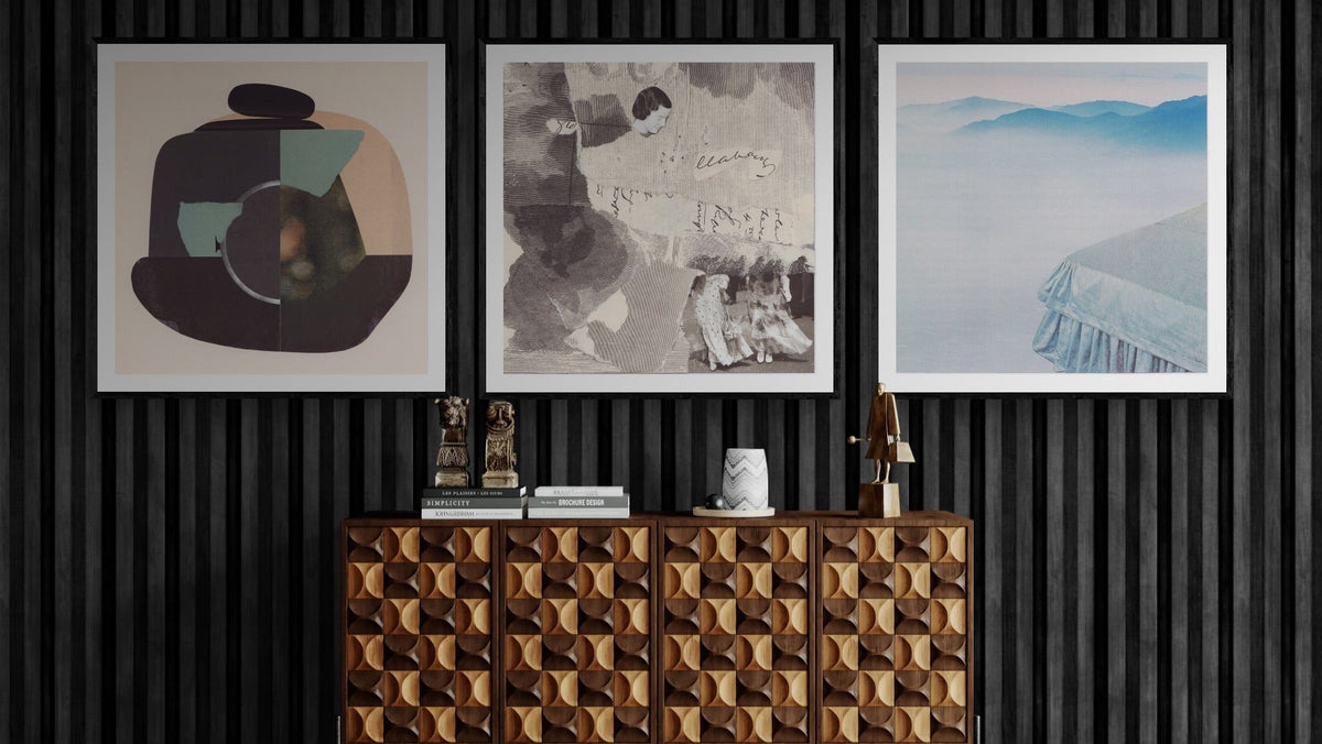 A styled interior scene showing three framed wall art prints hanging above a geometric wood sideboard against a black slatted wall. The left print is abstract with dark charcoal, beige, and teal shapes; the center print is monochrome with sketch-like figures and cursive writing; the right print shows a misty blue mountain landscape.
